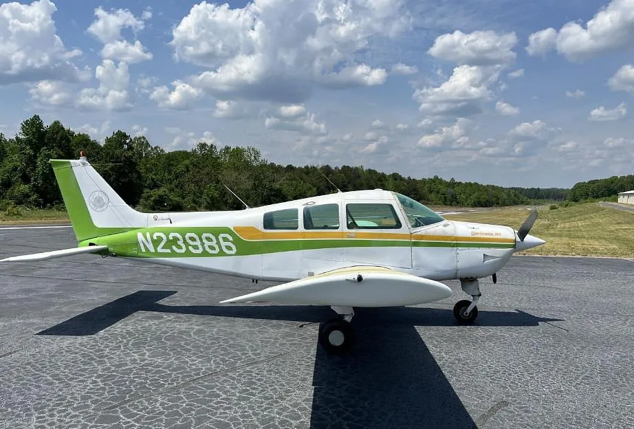 Side view of a white and green Beechcraft Sundowner 180 (N23986) on an airport tarmac with trees in the background.
