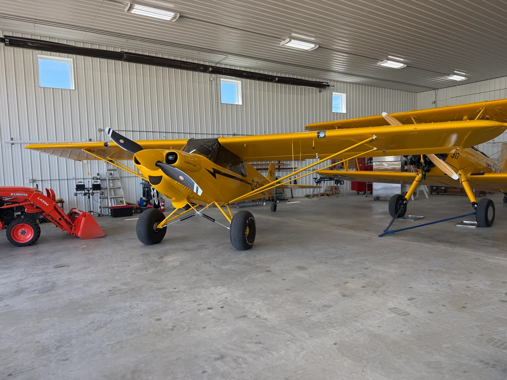 A yellow CubCrafters Carbon Cub EX-3 with large tires parked in a hangar, with another yellow aircraft nearby.