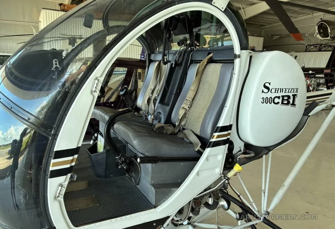 The two-seat interior cabin of a Schweizer 300CBi helicopter with gray seats and tan harnesses.