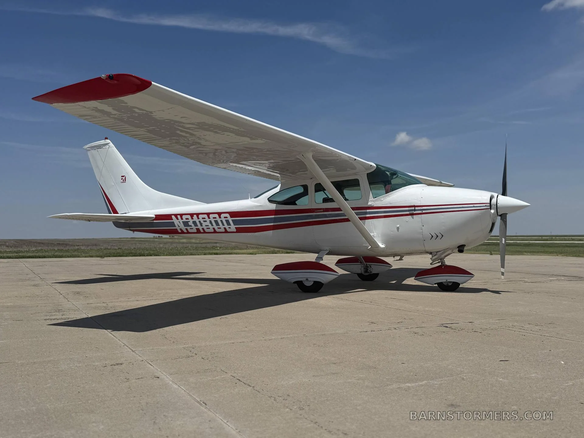 A white single-engine Cessna aircraft with red and grey stripes and red wingtips, parked on a concrete tarmac under a blue sky.
