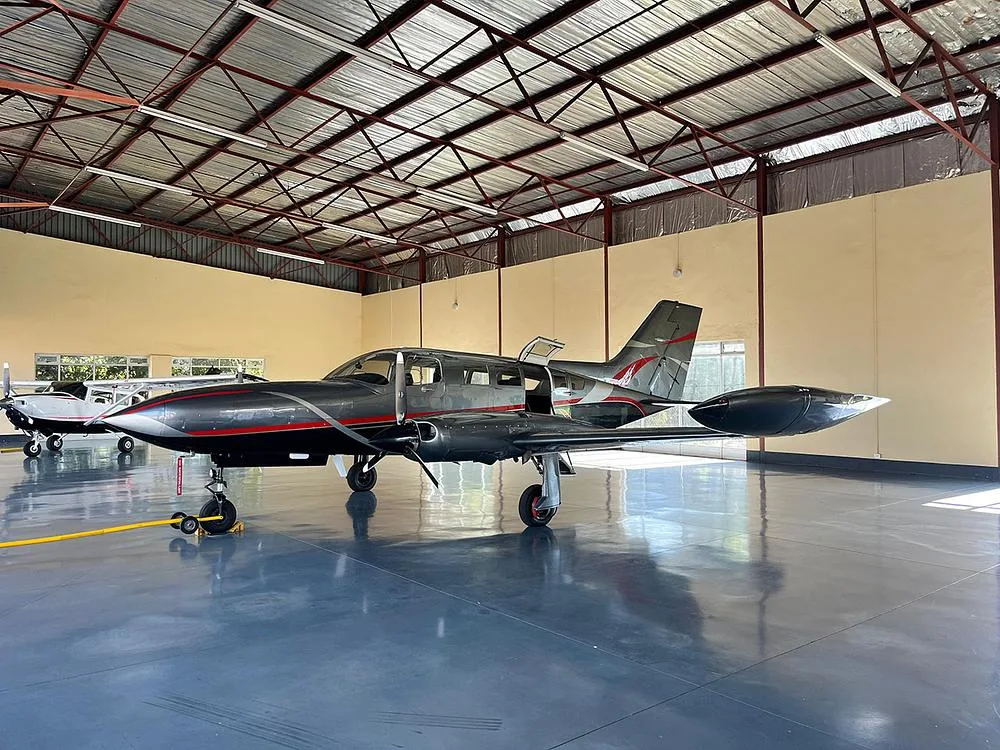 A dark grey twin-engine aircraft with red and silver accents, parked in a hangar with its passenger door open.