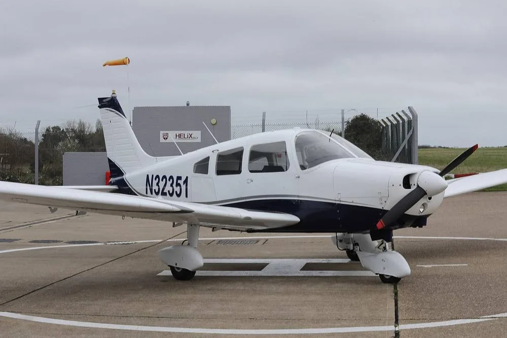 A white and blue Piper PA-28 aircraft with registration N32351 parked on an airfield with a windsock in the background.