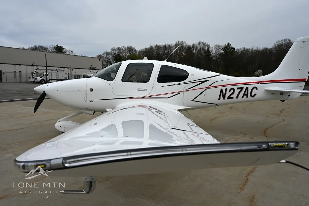 Close-up side view of a white Cirrus SR22 aircraft, tail number N27AC, showing the wing and fuselage.