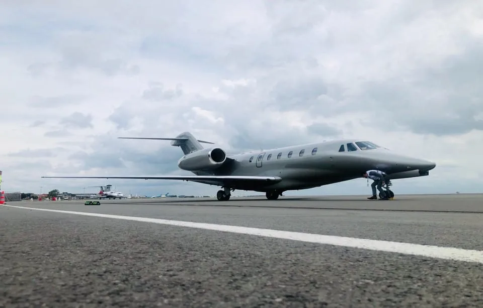 A silver private jet on an airfield with a person near the nose, under a cloudy sky.