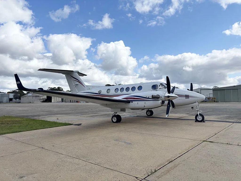 A white twin-turboprop aircraft with red and blue stripes, viewed from the side on a sunny day at an airport.