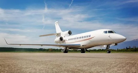 A white Dassault Falcon business jet with a red stripe parked on an airfield under a blue sky.