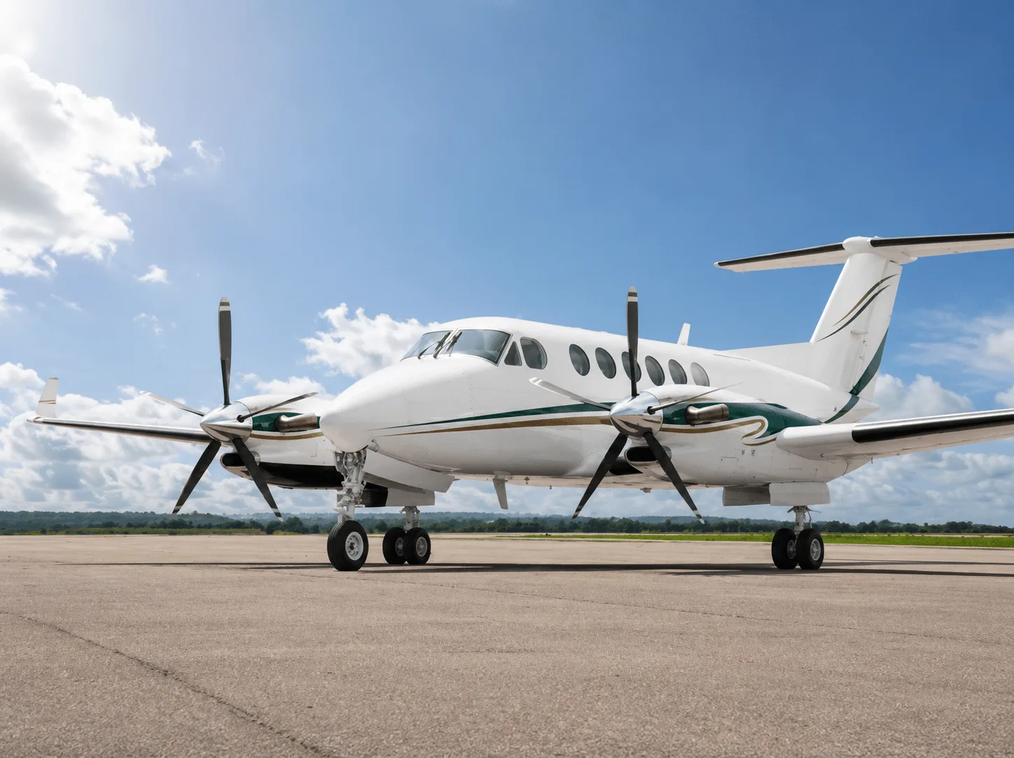 A white and green Beechcraft King Air turboprop aircraft is parked on an asphalt tarmac under a bright blue sky with scattered clouds.