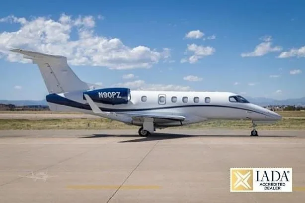 A white and blue Embraer Phenom 300 business jet, N90PZ, parked on an airfield under a blue sky.