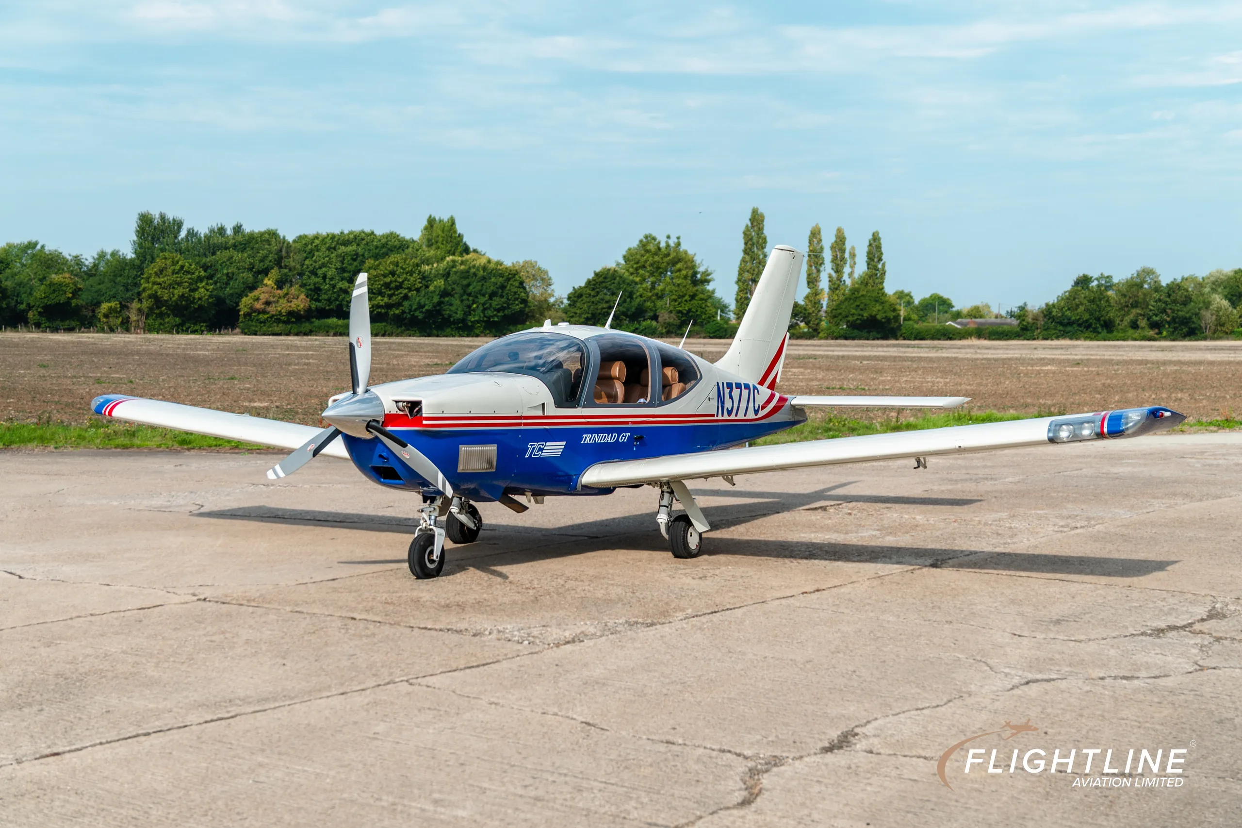 A front-left view of the N377C Socata Trinidad GT, painted white and blue with red stripes, parked on a concrete pad.