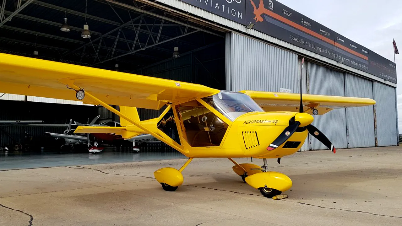 A yellow Aeroprakt-22 aircraft is parked on concrete in front of an open hangar with a company banner.