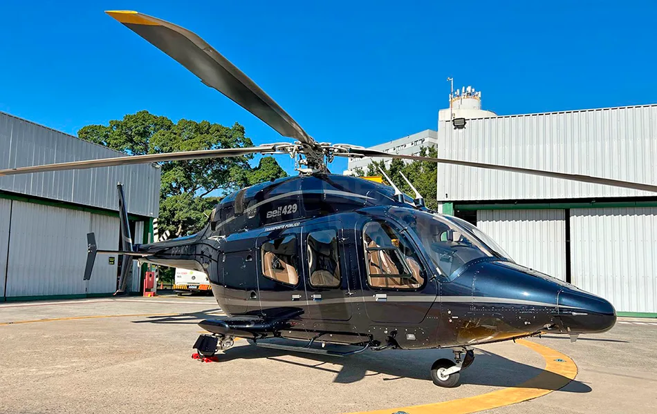A dark blue Bell 429 helicopter with tan interior parked on an airfield in front of a hangar under a clear blue sky.