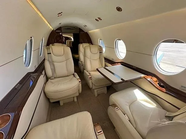 A view inside a private jet cabin shows light beige leather seats, a fold-out table, and dark wood trim.
