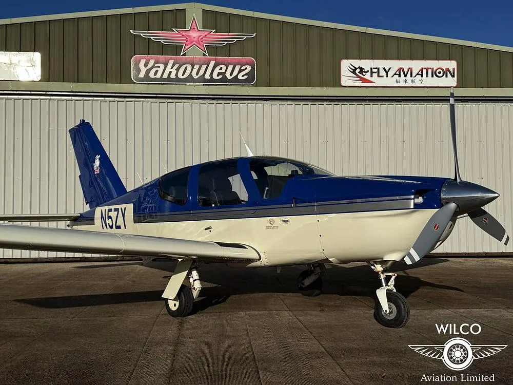 A blue and white single-engine aircraft, N5ZY, parked on an airfield in front of a hangar with 'Yakovlev' and 'Fly Aviation' signs.