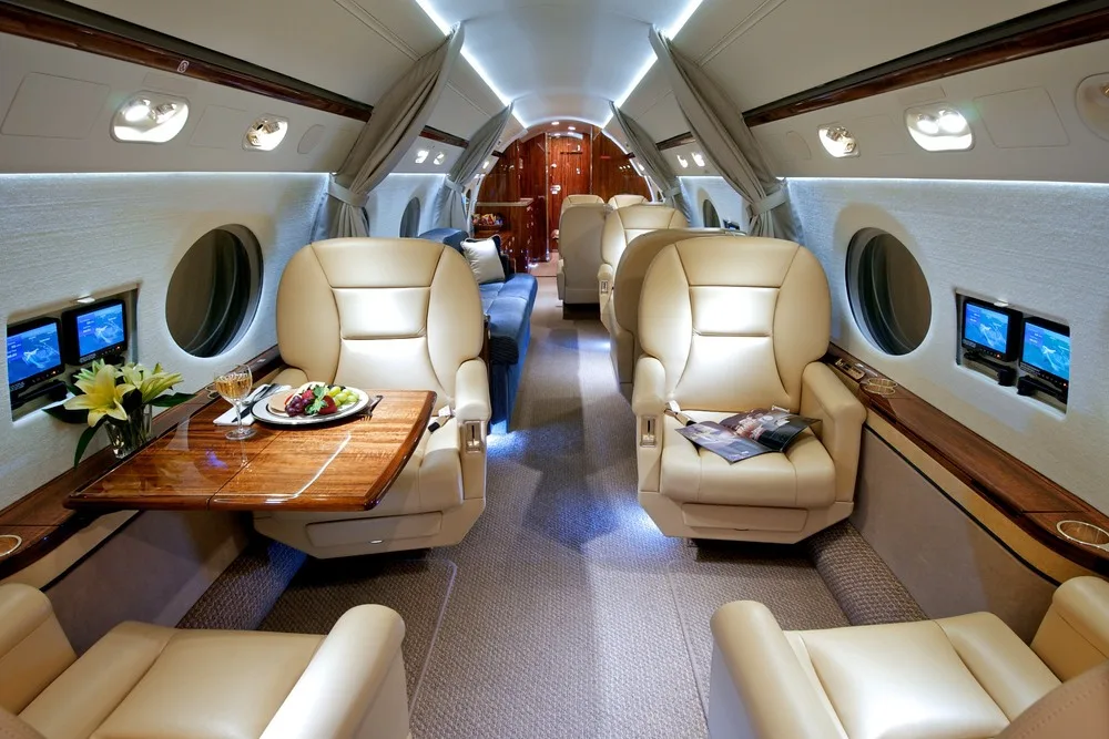 The luxurious cabin interior of a private jet, featuring tan leather seats, a wooden table with a meal, and individual entertainment screens.