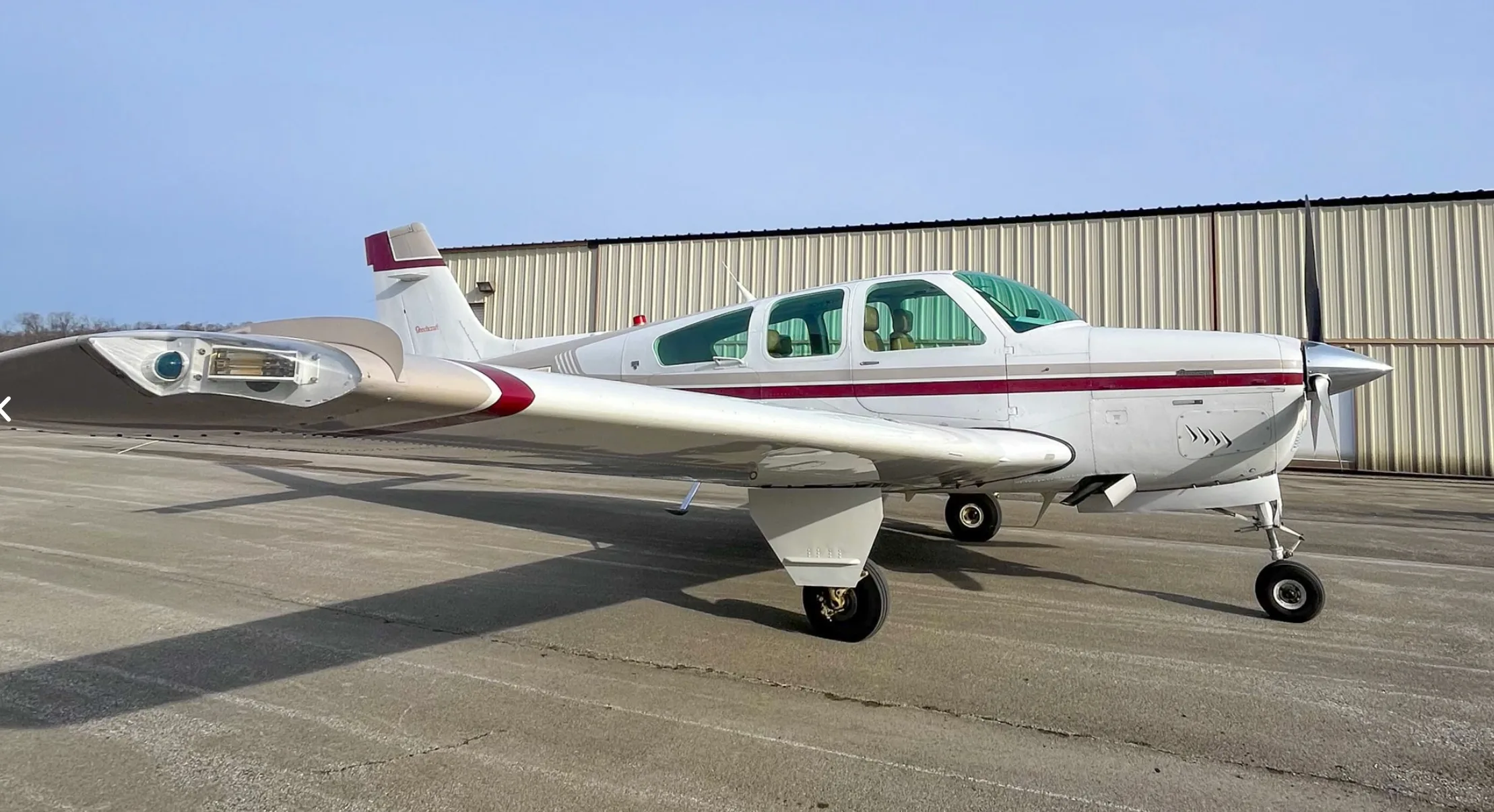 A white and maroon Beechcraft Bonanza aircraft is parked on a tarmac with a hangar in the background.