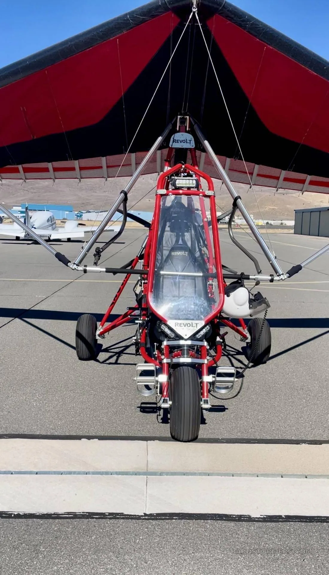 A red and black trike ultralight aircraft, branded 'REVOLT', viewed head-on while parked on an airport tarmac.