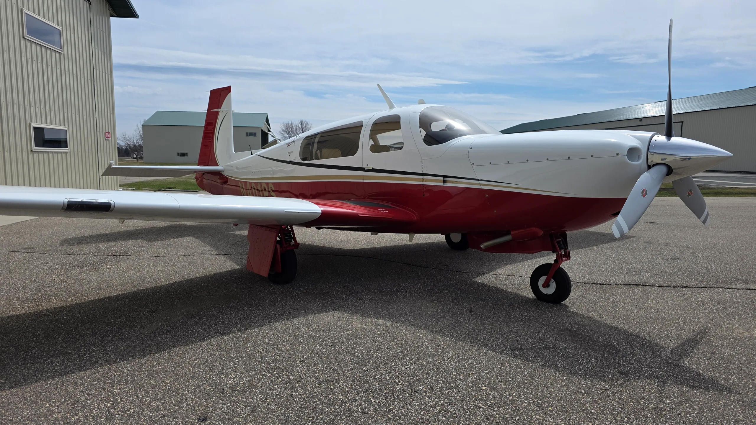 A red and white Mooney aircraft with gold stripes parked on an asphalt apron, viewed from the front right.