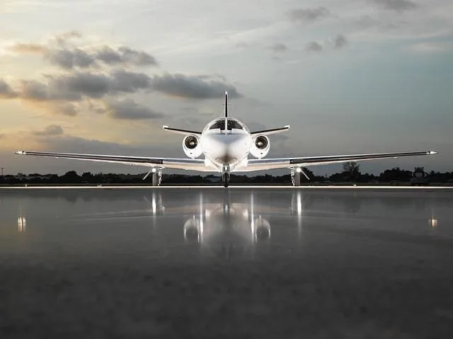 A white Cessna Citation CJ4 is viewed head-on on a tarmac at dusk, with reflections on the ground.