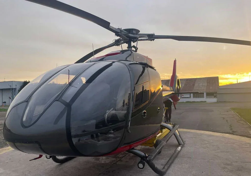 A dark grey and red helicopter sits on a helipad at sunset, viewed from the front-left.