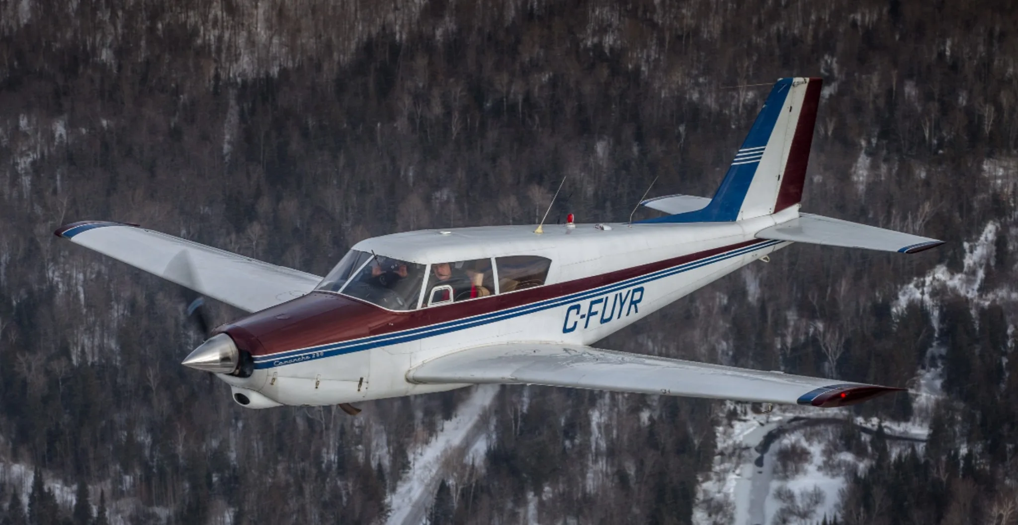A white, maroon, and blue Piper Comanche 260 aircraft (C-FUYR) flying over a snowy, forested landscape.
