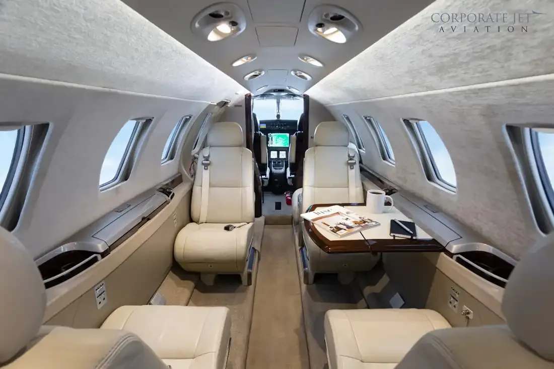 The interior of a private jet cabin with light-colored leather seats and a table with a magazine and coffee cup.
