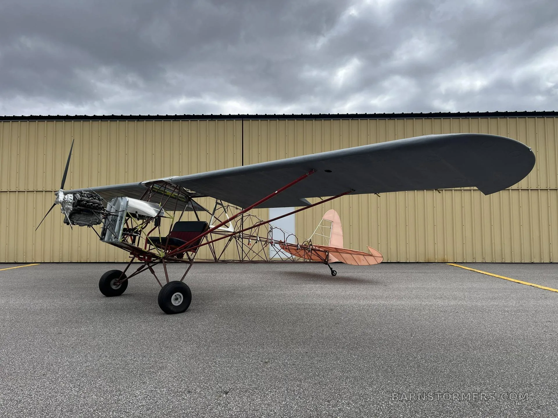 A side view of an experimental ultralight aircraft with an exposed frame, a single wing, and a propeller, parked on an asphalt apron in front of a yellow hangar.