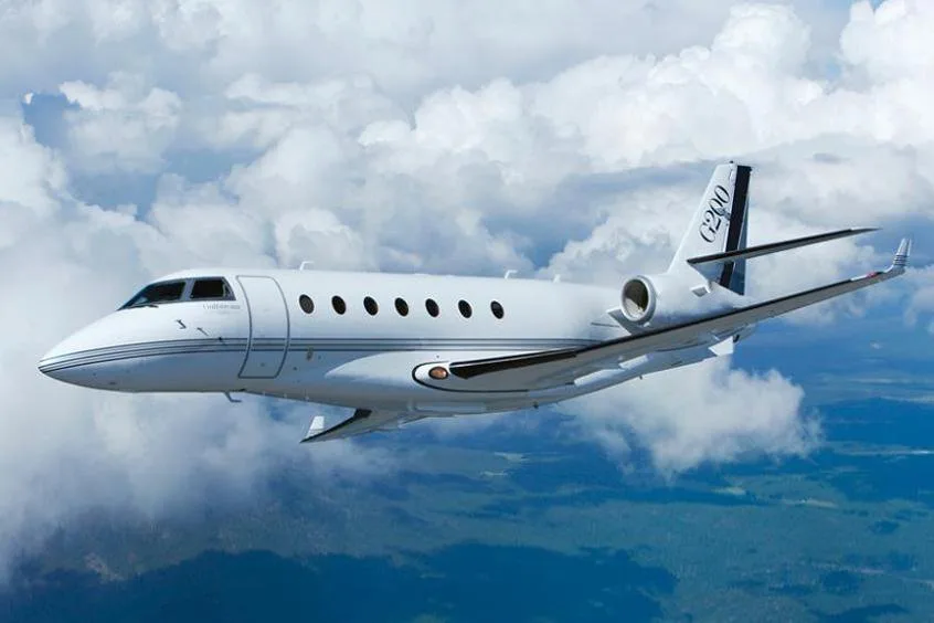 A white Gulfstream G200 business jet with black tail stripes flies through a cloudy sky over green terrain.