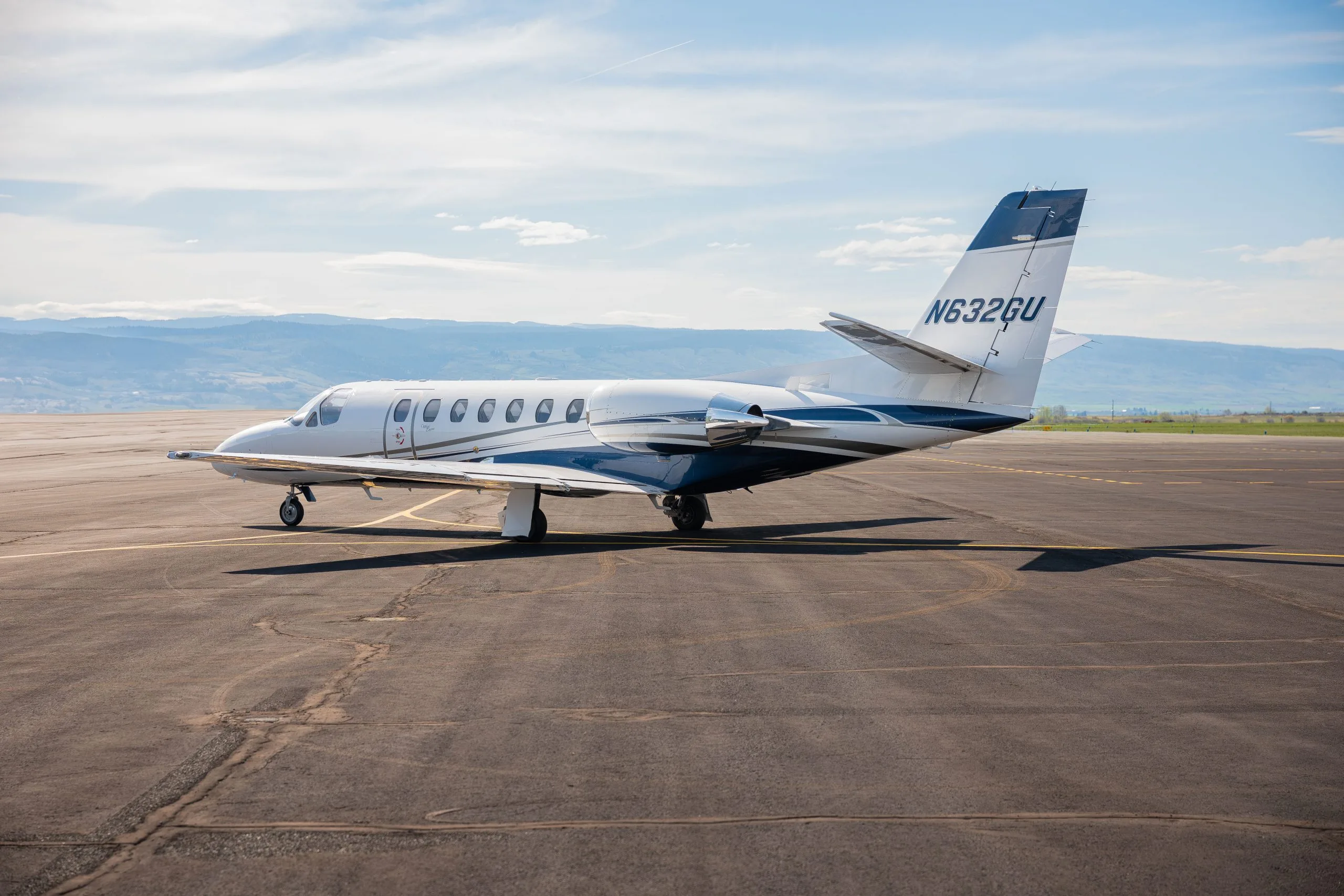 A white Cessna Citation business jet with blue and gray stripes, parked on an airfield with distant mountains under a bright sky.