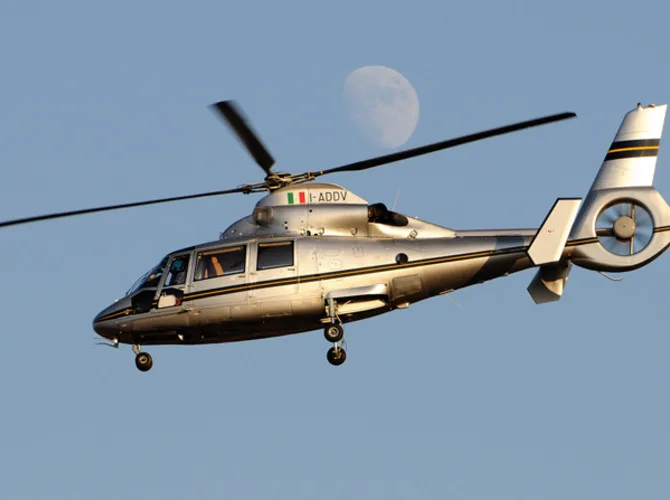 A silver helicopter with registration I-ADDV flies against a blue sky with the moon visible in the background.
