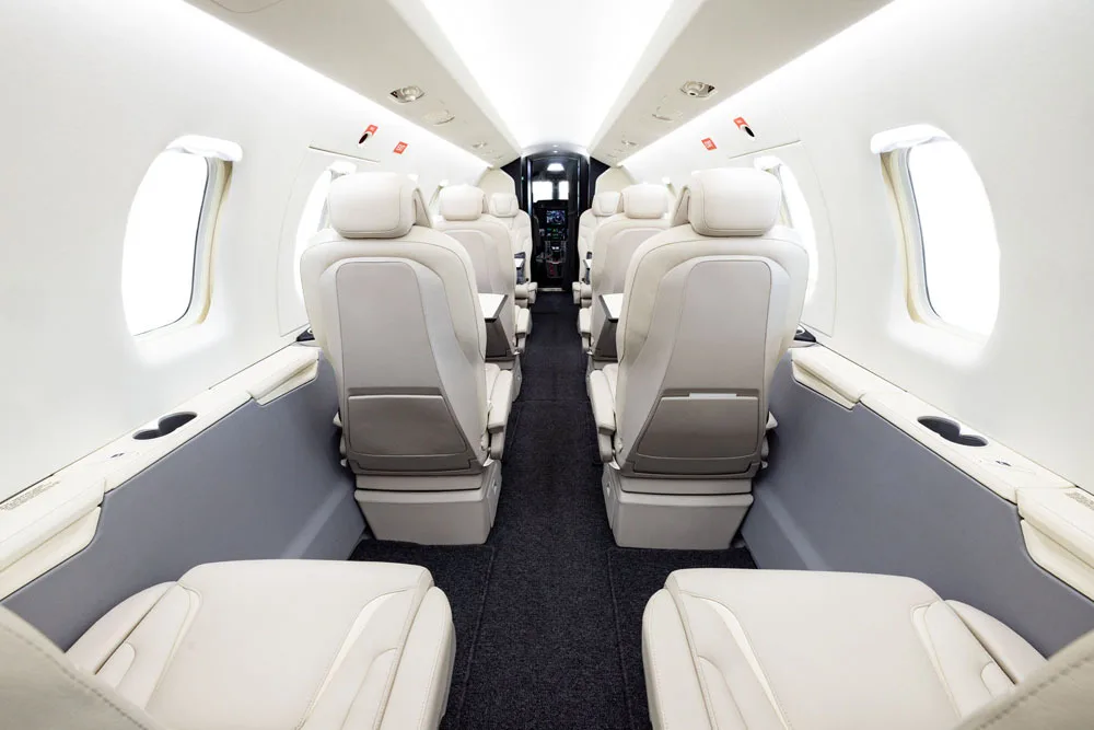 A wide-angle view of a private jet cabin with multiple rows of light beige seats and a dark aisle carpet.