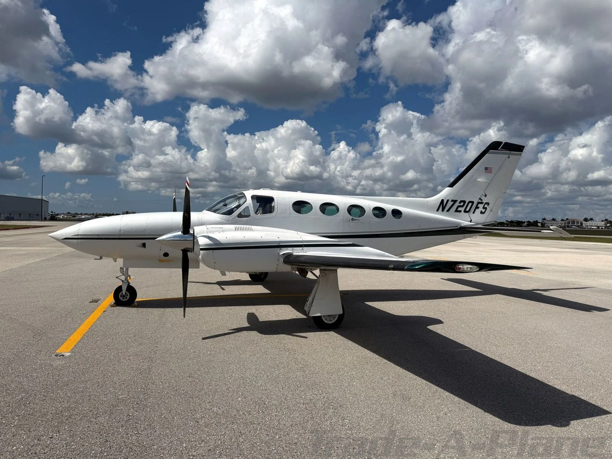 A white Cessna 414 Chancellor aircraft, N720FS, with dark green stripes, parked on an airfield under a partly cloudy sky, viewed from the front left.