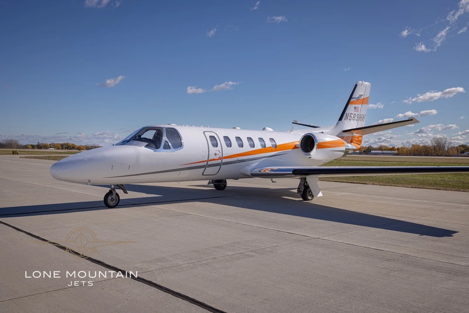 A white Cessna Citation Bravo business jet with orange and grey stripes on the tarmac under a blue sky.