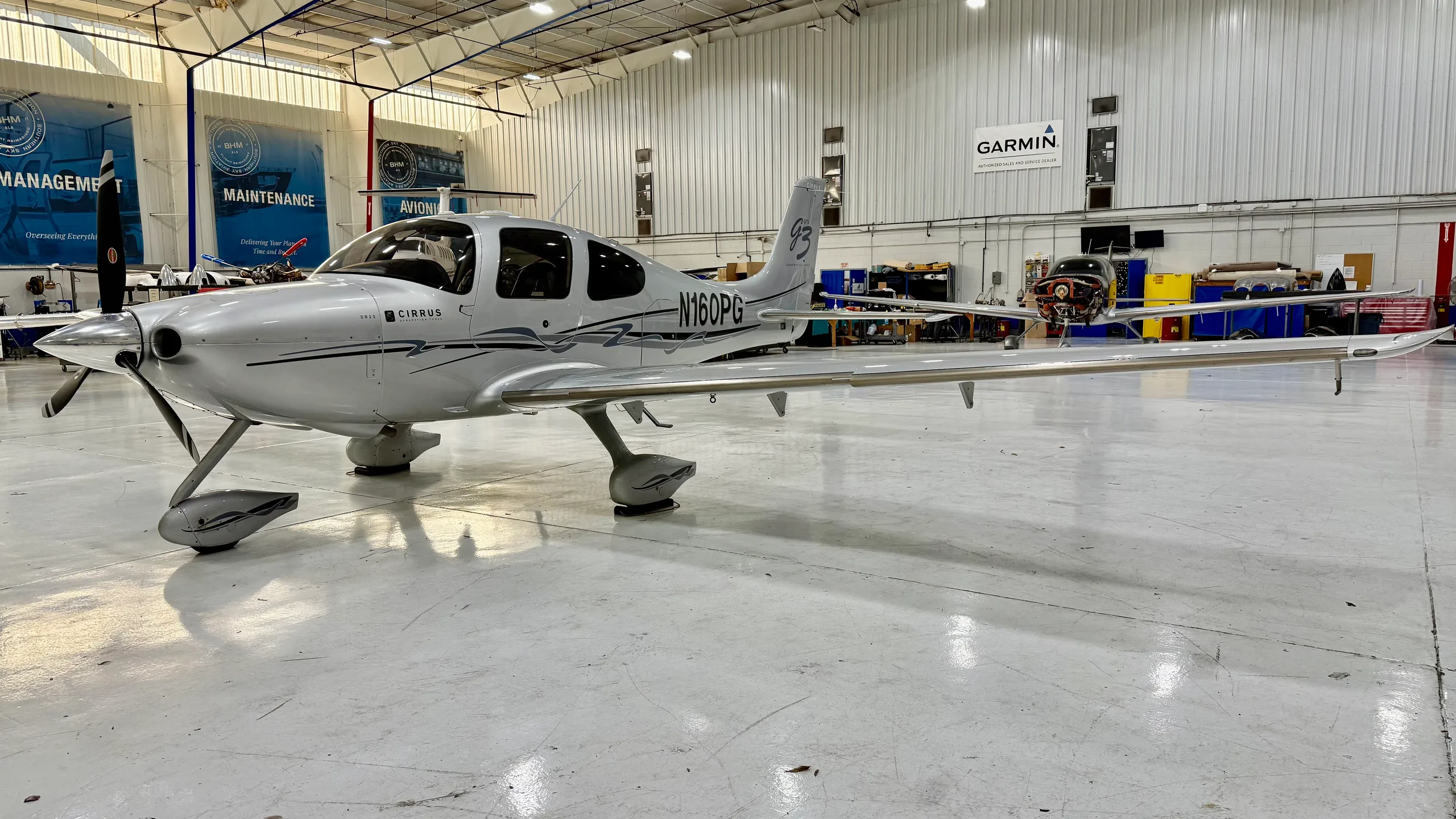 A silver Cirrus SR22 aircraft with dark grey accents, viewed from the front-left in a hangar.
