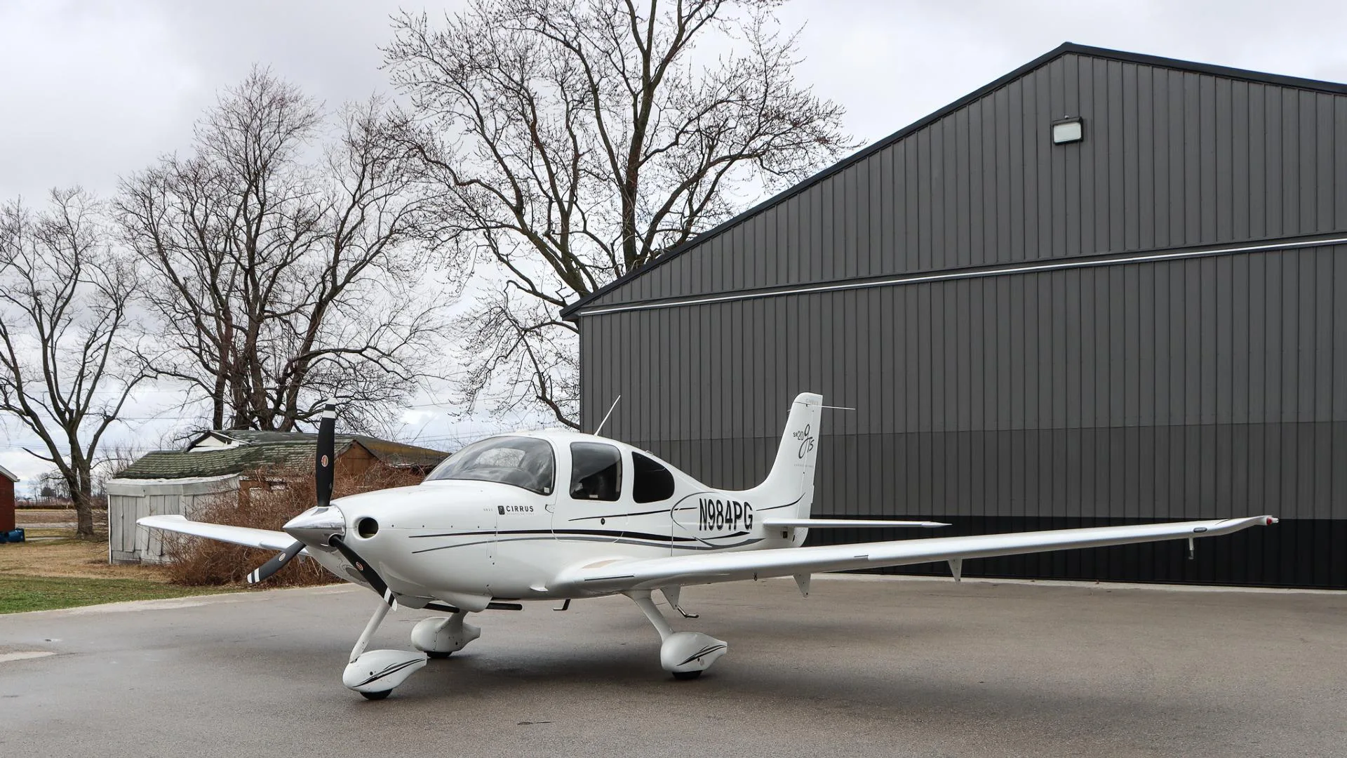 A white Cirrus SR20 GTS aircraft, N984PG, is parked with its right side visible in front of a dark gray hangar and bare trees.