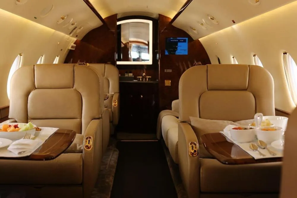 The private jet's interior with two beige seats, tables with food, and a lavatory entrance in the background.