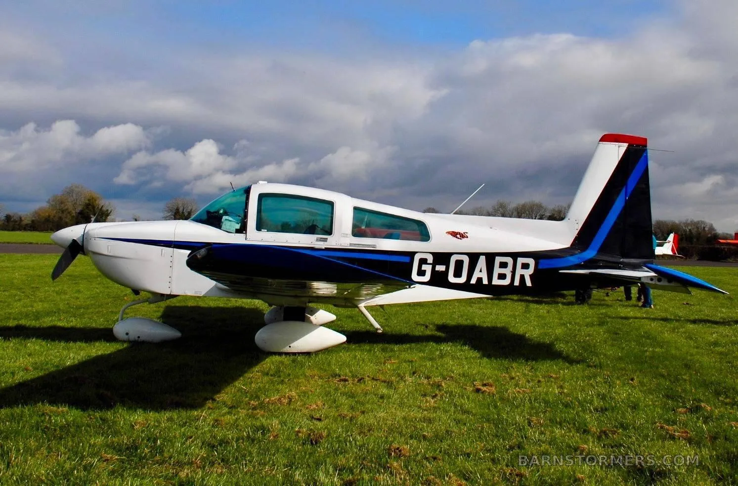 A white and blue light aircraft, registration G-OABR, parked on a green grassy field under a partly cloudy sky.
