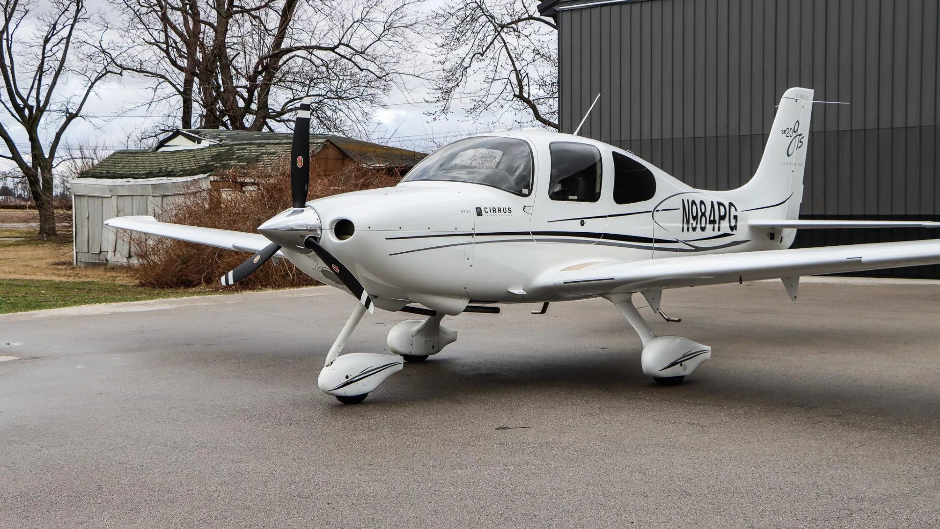 A white Cirrus SR20 GTS aircraft, N984PG, is parked with its right side visible against a dark gray hangar.