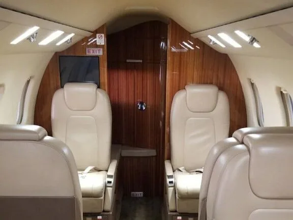 The interior cabin of a private jet with light beige leather seats and wood paneling, showing an "EXIT" sign above the door.