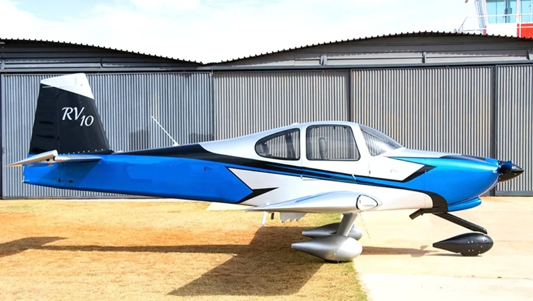 A blue, silver, and black Van's RV-10 aircraft parked on a grassy patch in front of a hangar.