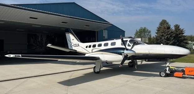 A white and dark blue Cessna 414 Chancellor twin-engine aircraft with gold stripes parked on an apron in front of a blue hangar.