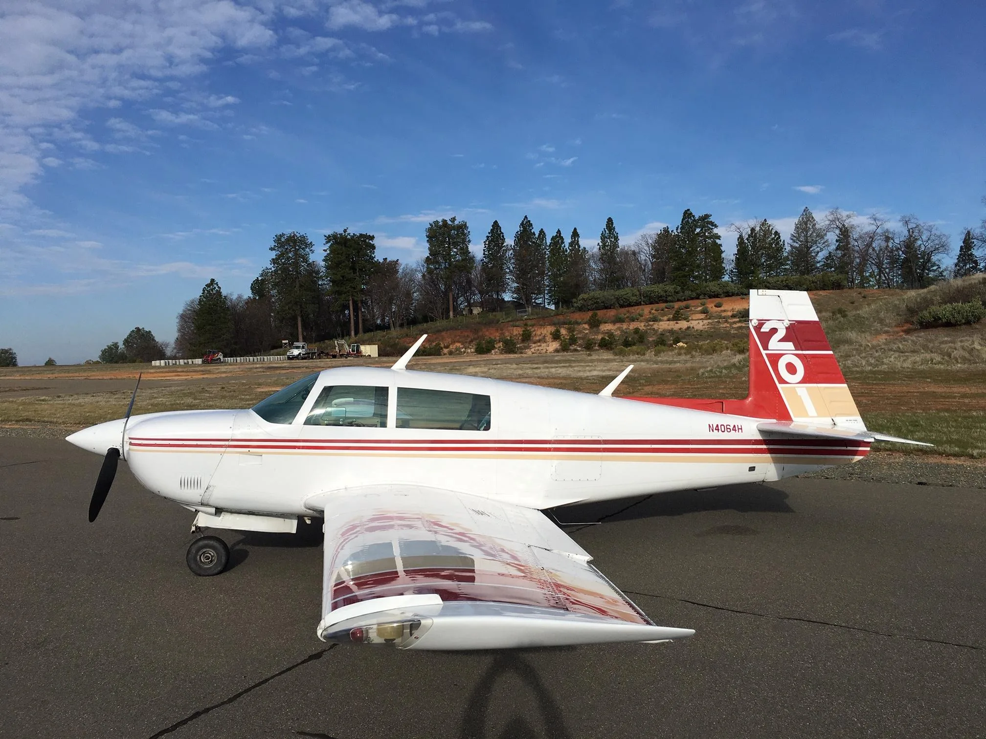 A white Mooney M20 aircraft with red and gold stripes and a red '20' tail, parked on an asphalt surface with trees and a hill in the background under a blue sky.