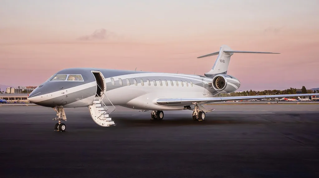 A Bombardier Global 7500 private jet with a grey and white paint scheme parked on a tarmac at sunset, with its main door open and stairs extended.
