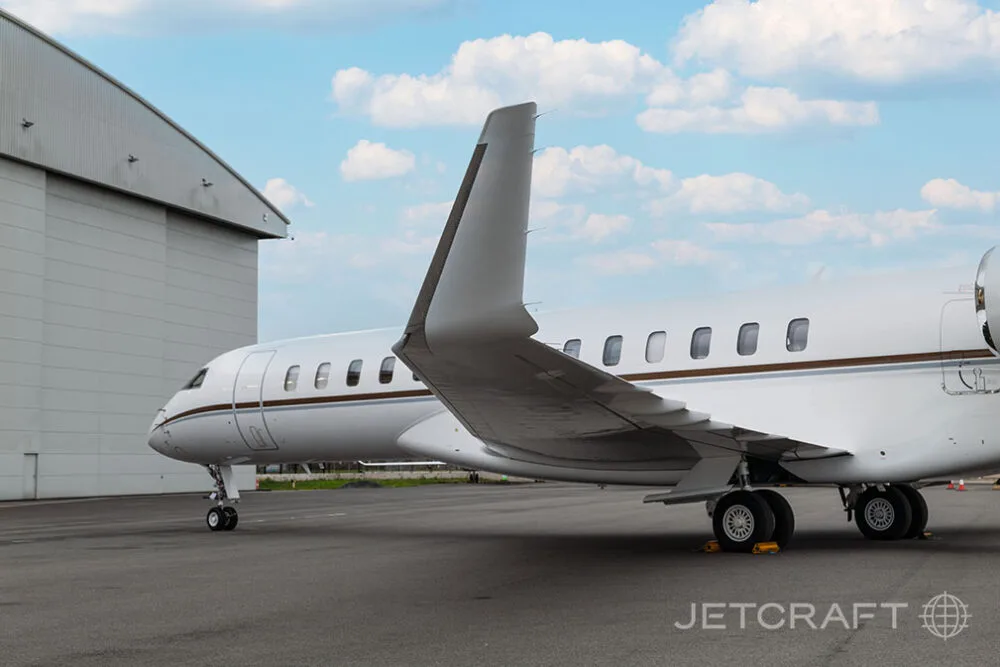 The rear fuselage and tail of a white Bombardier Global business jet parked next to a large hangar.