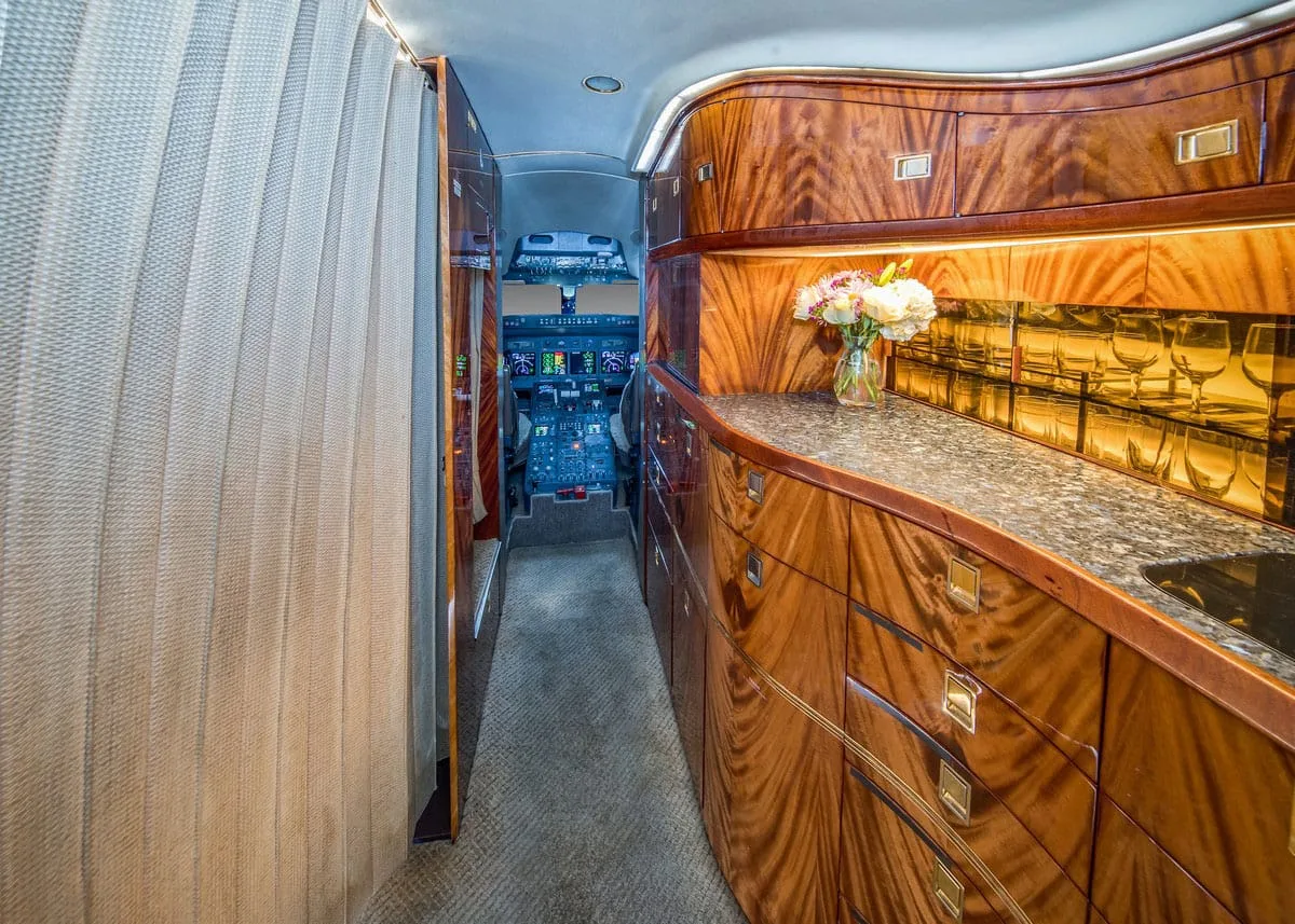 A view down the aircraft aisle from the galley, featuring polished wooden cabinets, a granite countertop with flowers, and the cockpit visible in the distance.