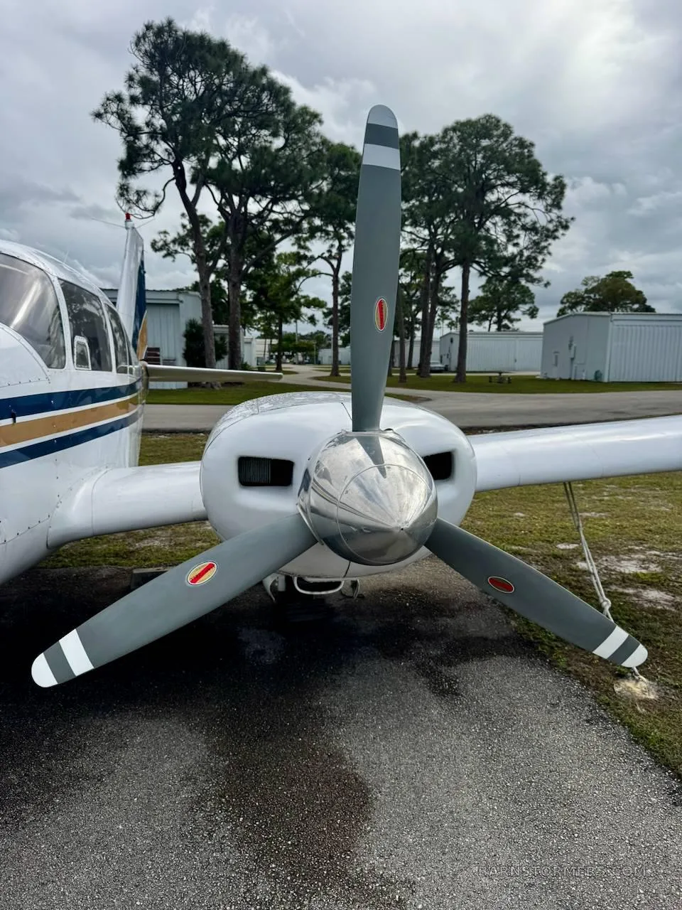 A close-up front view of the propeller and engine cowling of a white twin-engine aircraft, with trees and hangars in the background.