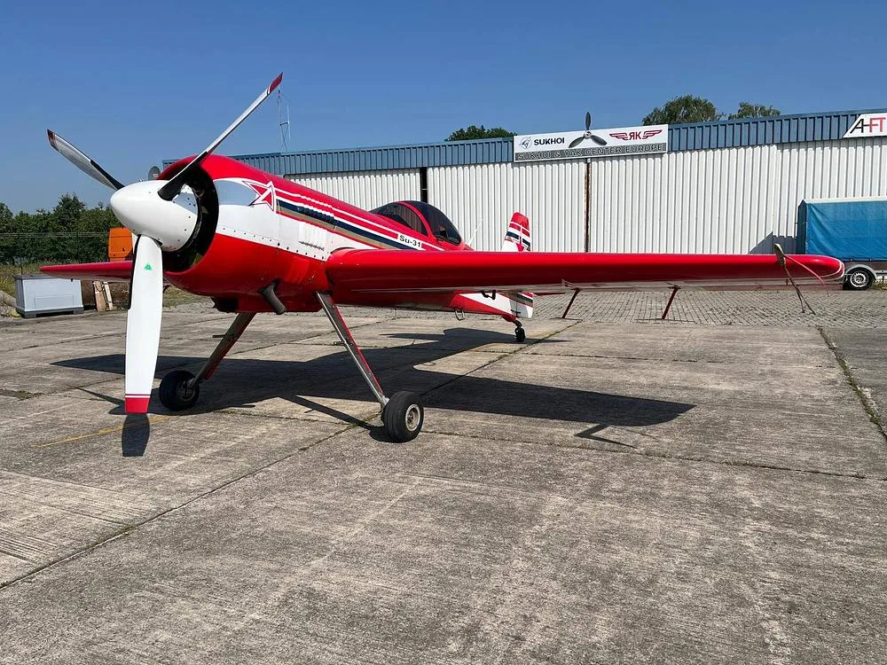 A red and white Sukhoi Su-31 aerobatic aircraft with a white propeller is parked on a concrete tarmac, with a hangar in the background.