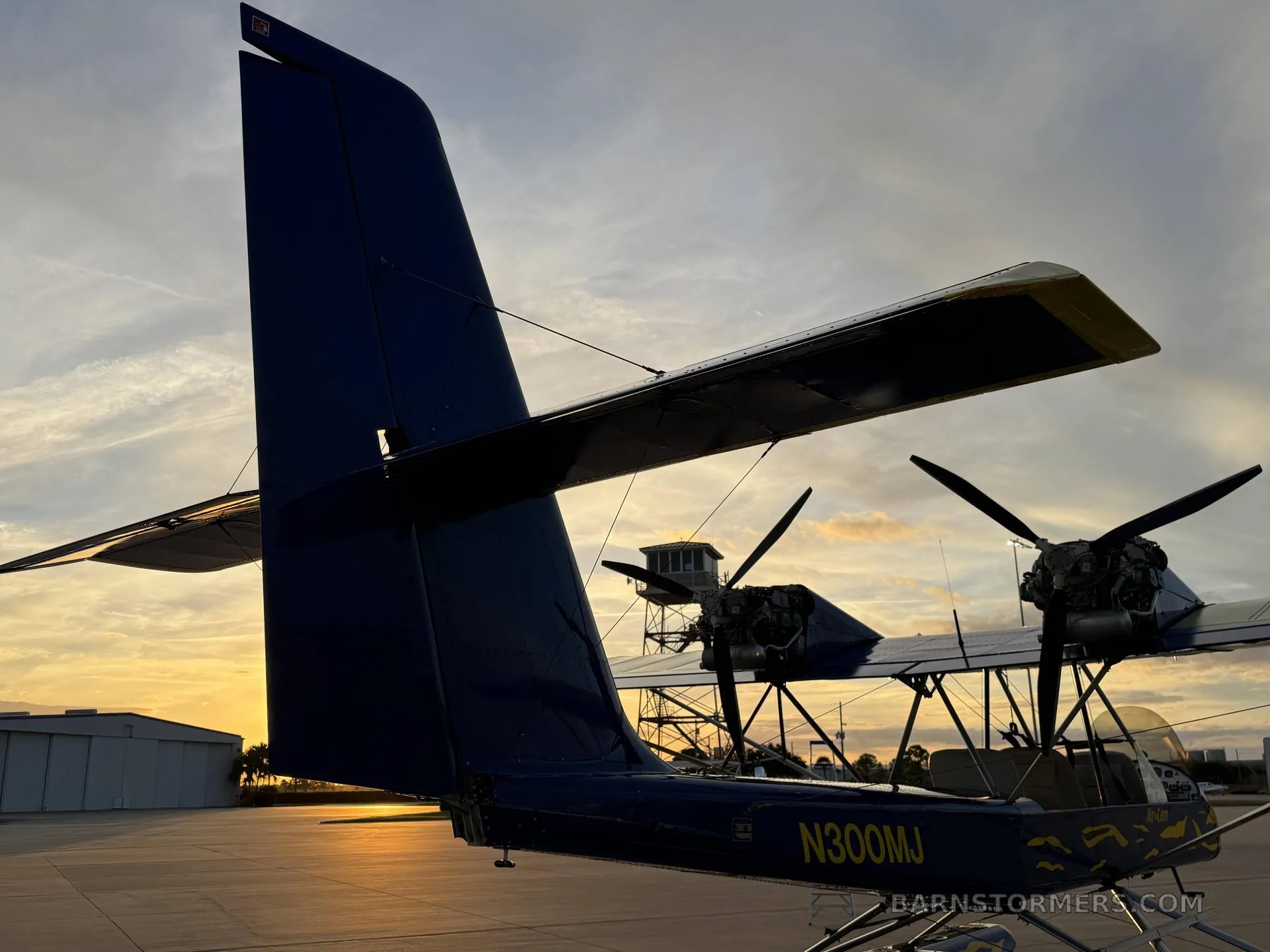 A rear view of the blue and yellow Air-Cam seaplane, showing the tail and engines against a sunset sky with a control tower in the background.