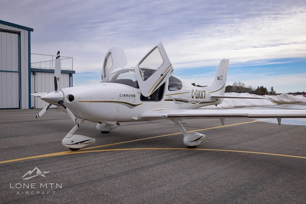 A white Cirrus SR22 aircraft with registration C-GKKT parked on an airfield with its doors open, a hangar in the background.