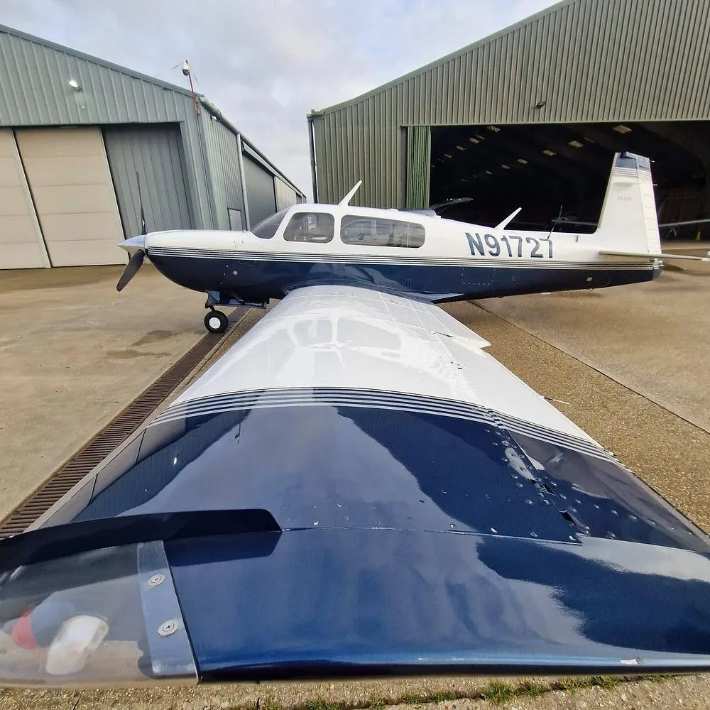 A white and blue single-engine aircraft, N91727, is parked on an airfield next to hangars, viewed from over its wing.