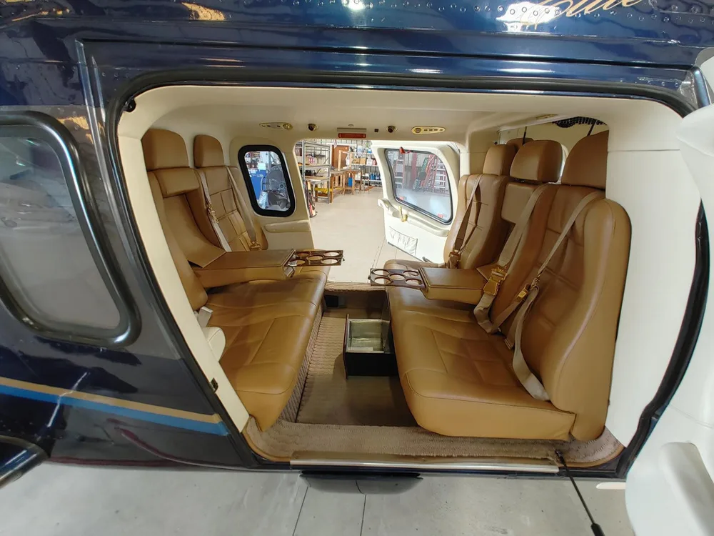 The spacious tan leather interior cabin of a helicopter shows two rows of seats with cup holders and open doors.